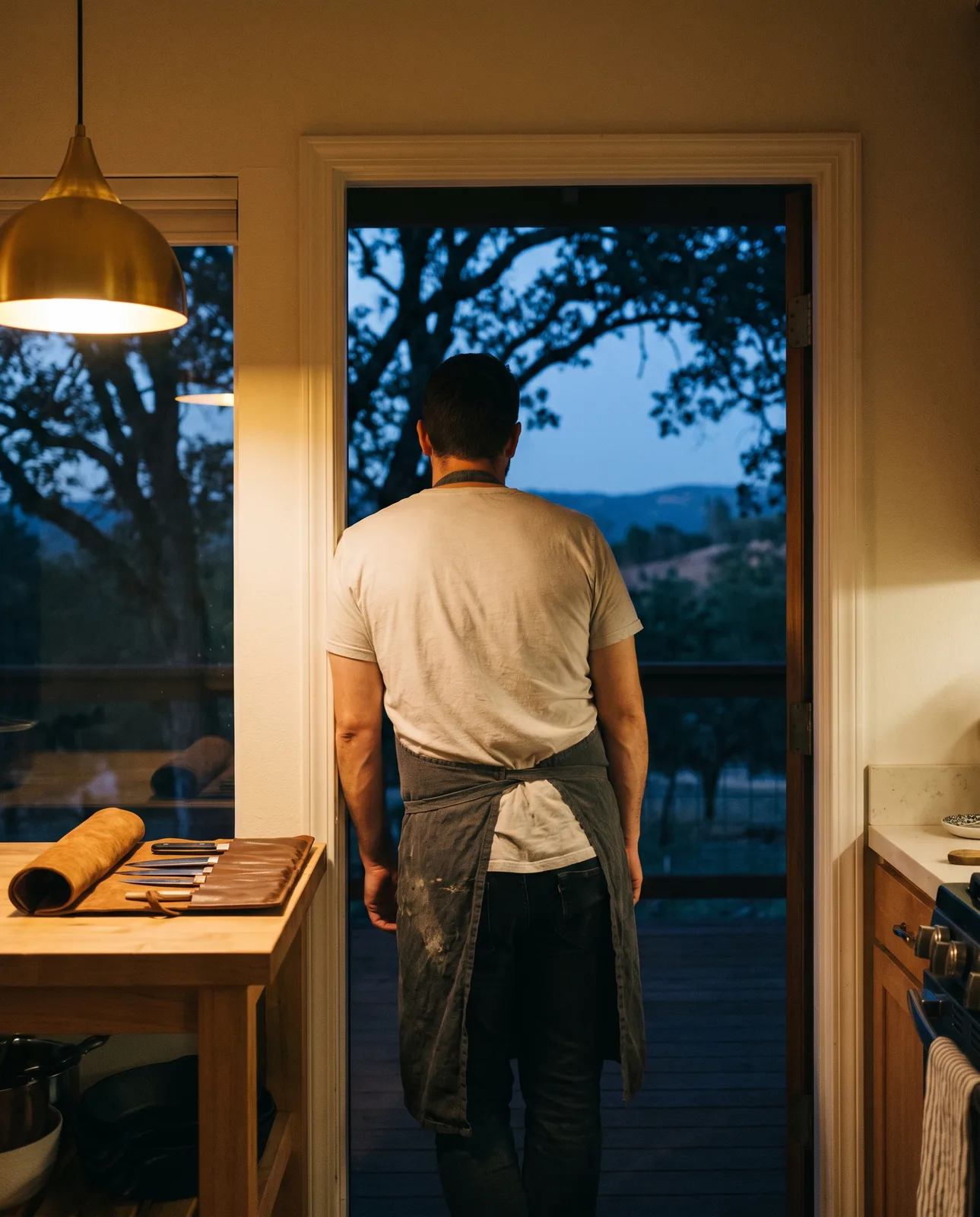 A chef in a charcoal apron at the open back door of a home kitchen at dusk, looking out at oaks and a Foothills hillside