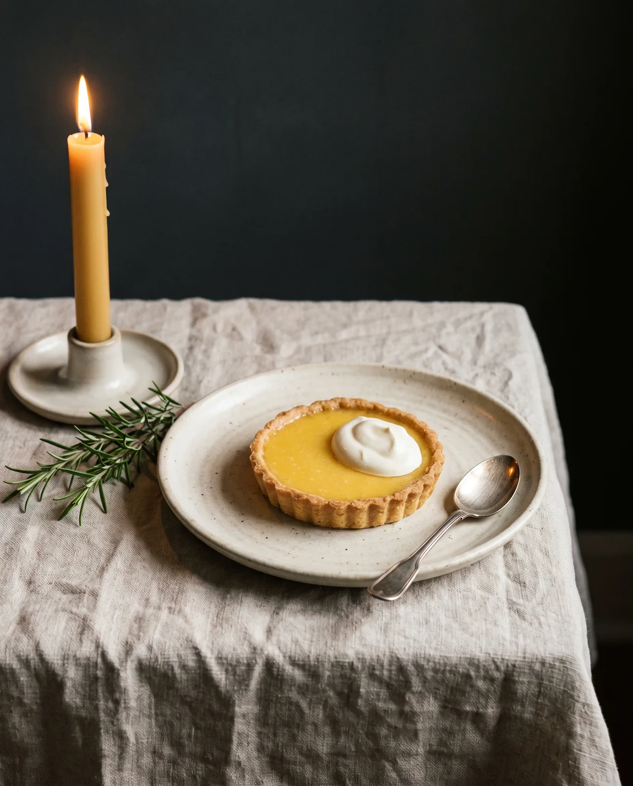 A lemon tart with crème fraîche on a cream plate beside a lit taper candle